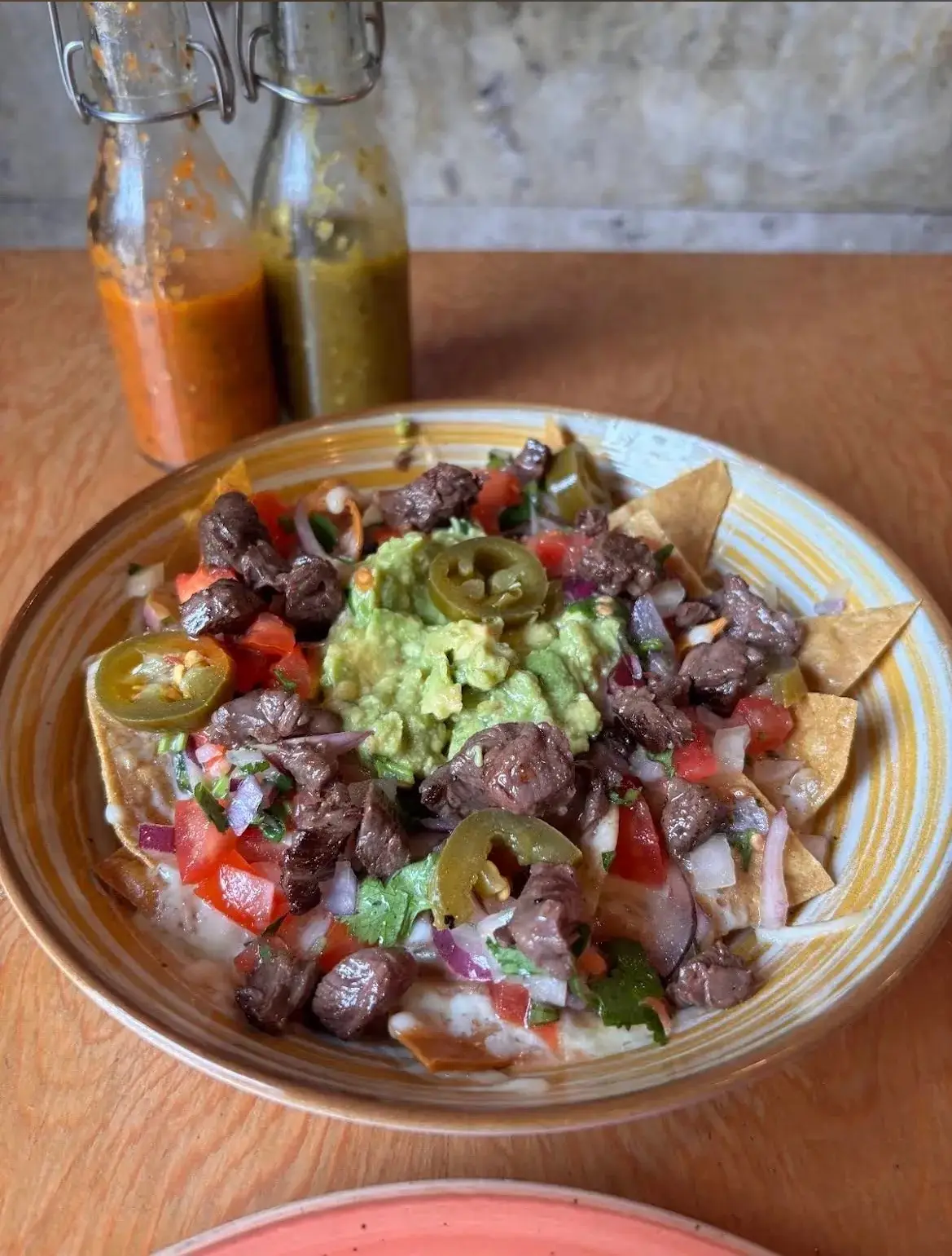 Loaded nachos with steak, guacamole, pico de gallo, red onion, and pickled jalapeños on a ceramic plate, with house hot sauces in the background, at Taquerías Tamarindo