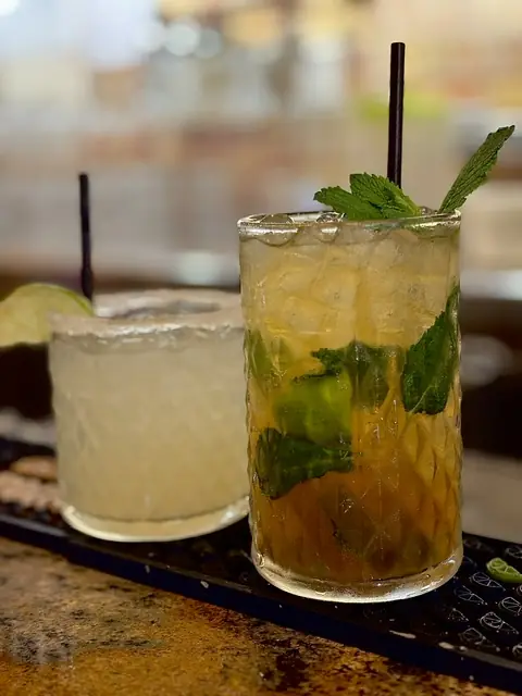 Two cocktails on a bar counter, one pale yellow with a lime wedge and one amber with fresh mint leaves and ice, at Taquerías Tamarindo