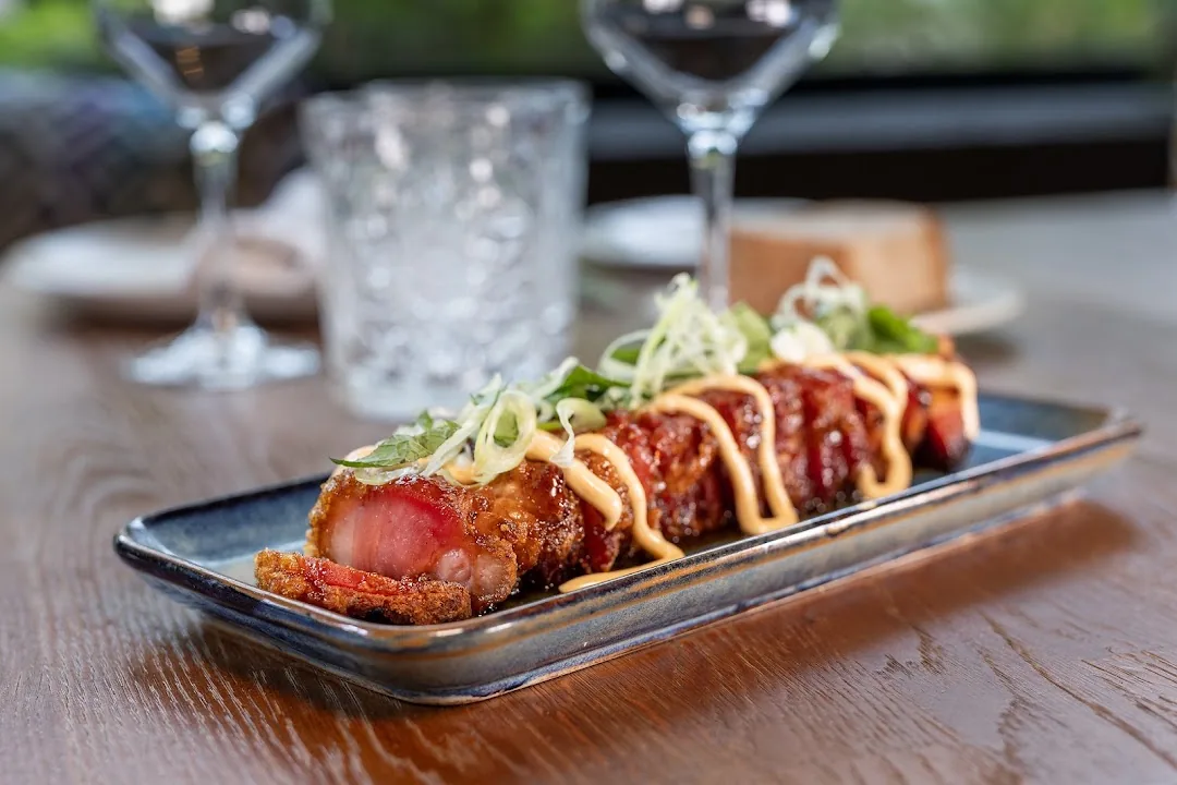 A crispy glazed pork dish topped with sliced scallion and herbs, drizzled with a creamy sauce on a dark rectangular plate