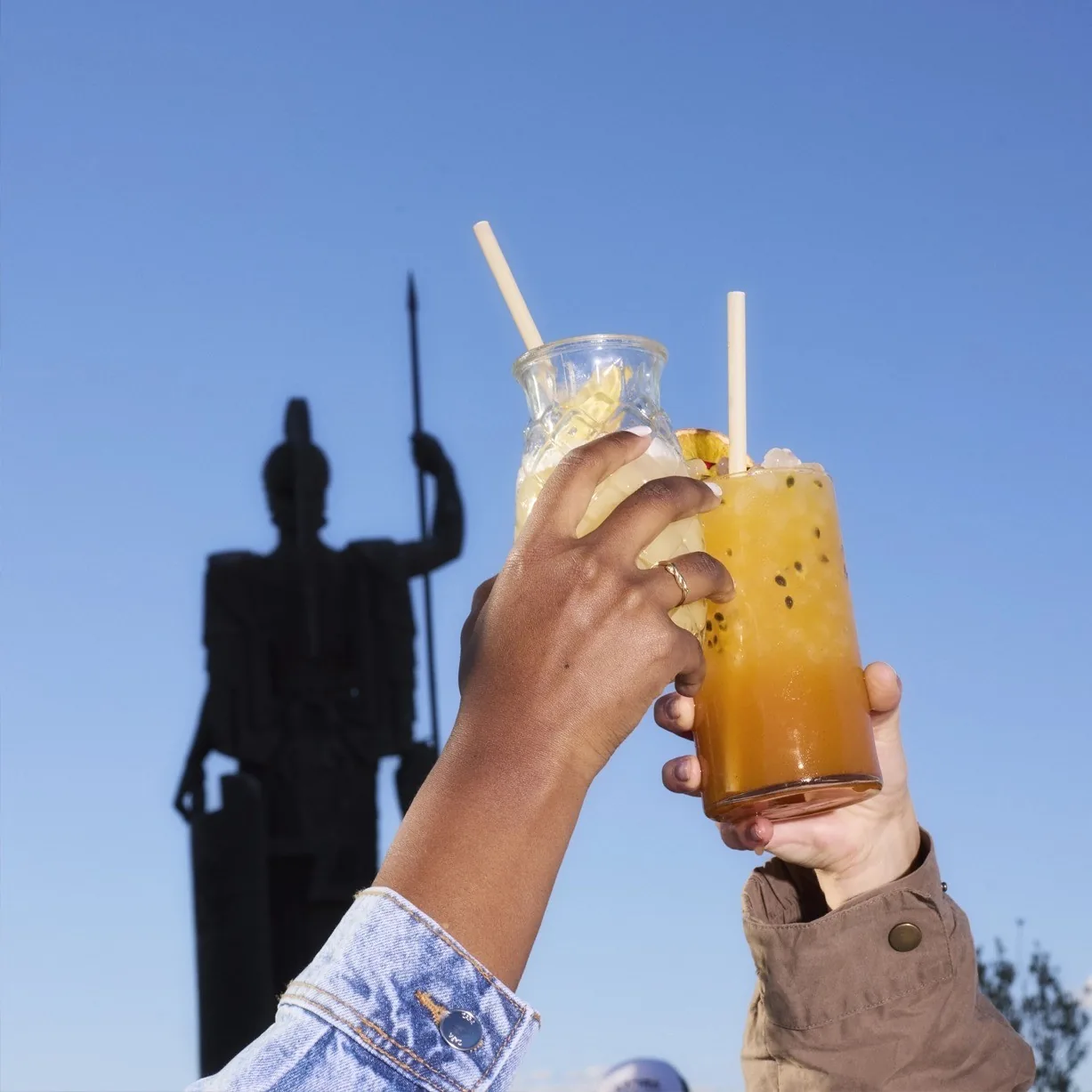 A hand holding up an orange cocktail with straws against a blue sky and a statue silhouette