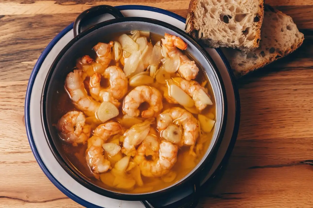 Blue-rimmed cazuela of gambas al ajillo, prawns poaching in golden garlic oil with sliced garlic, two slices of rustic bread beside it on a wooden table