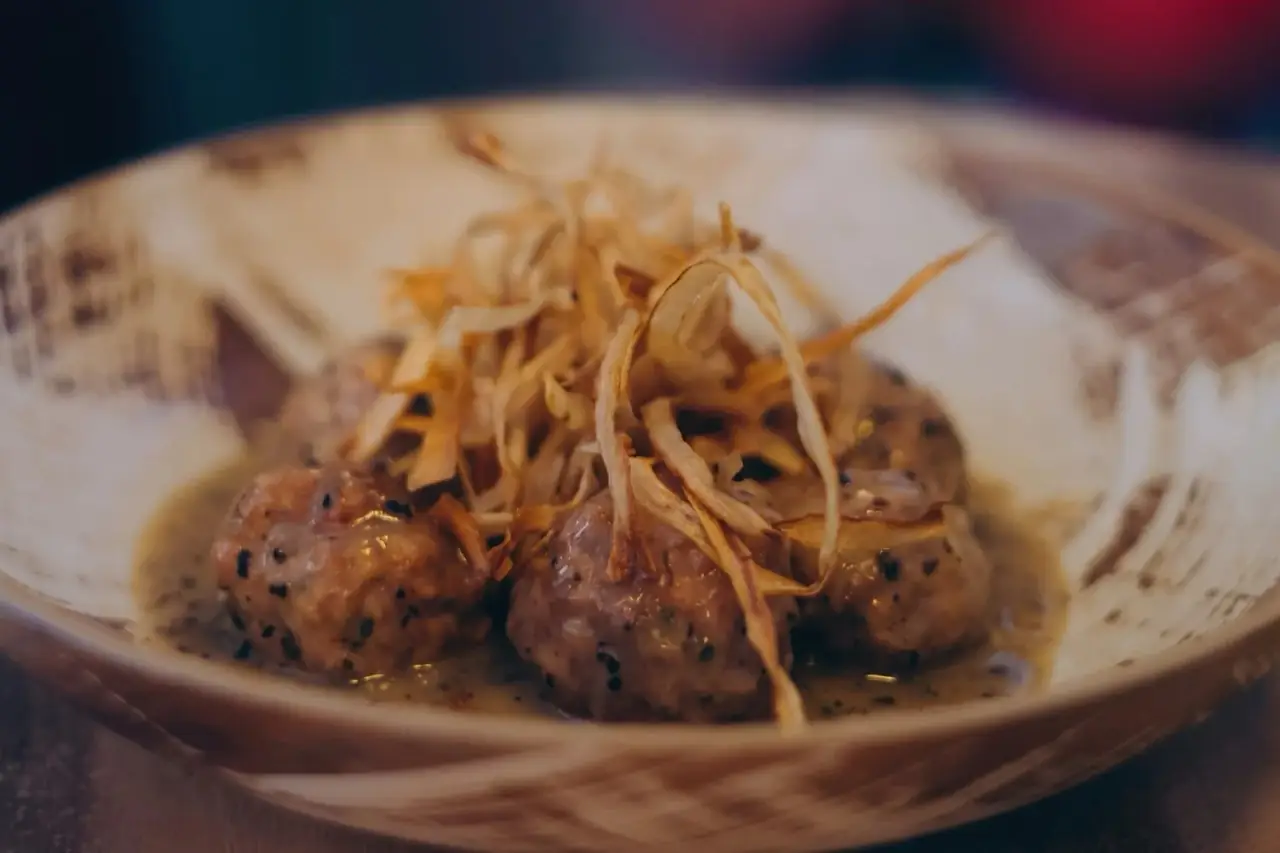 Three meatballs in a pale creamy sauce topped with crispy fried leek strands, served in a rustic ceramic bowl