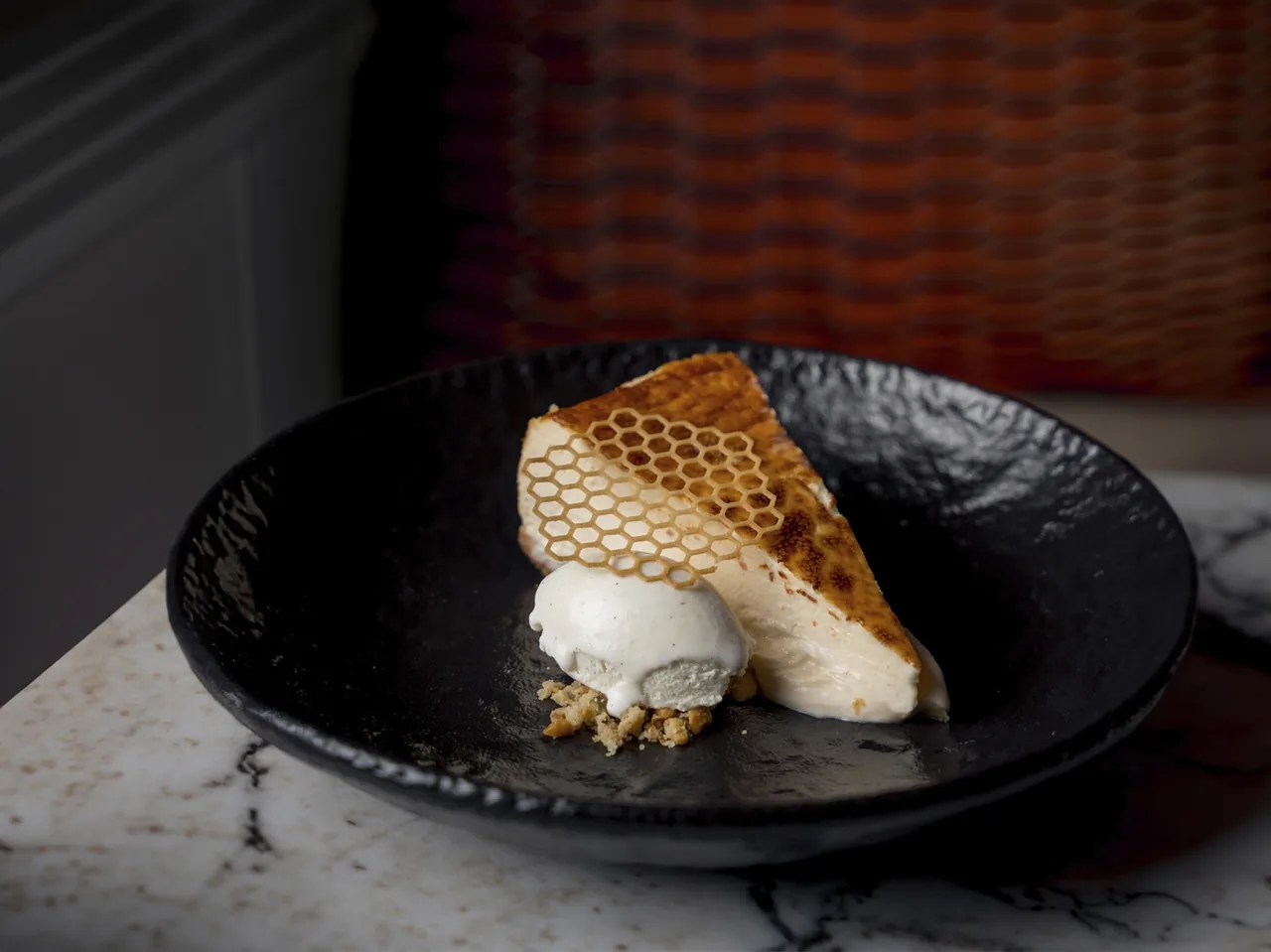 A slice of cheesecake with a browned top and a honeycomb-patterned tuile beside a scoop of ice cream on a black bowl
