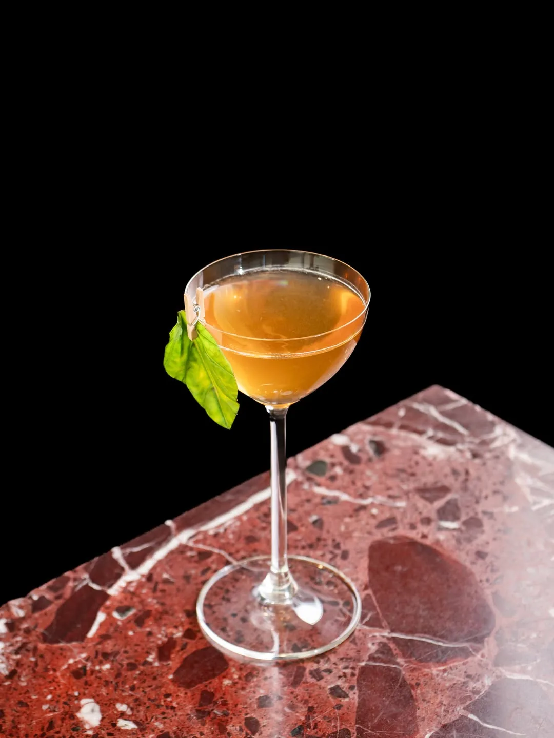 An orange-hued cocktail in a coupe glass garnished with a basil leaf, on a marble surface