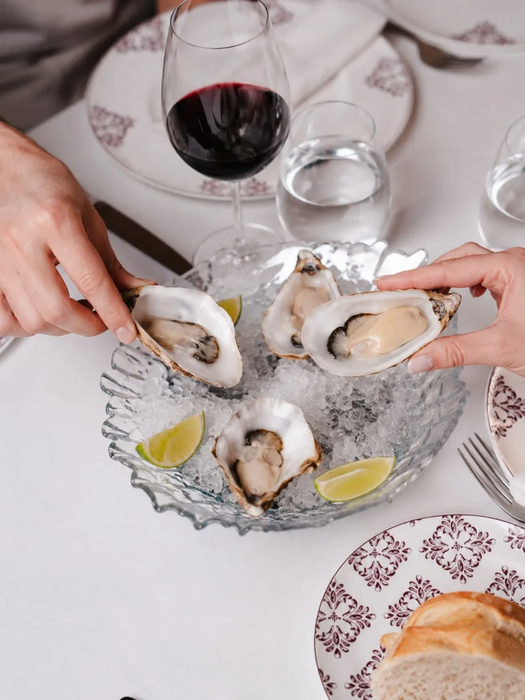 A silver platter of fresh oysters on ice with lemon wedges, next to glasses of red wine and bread