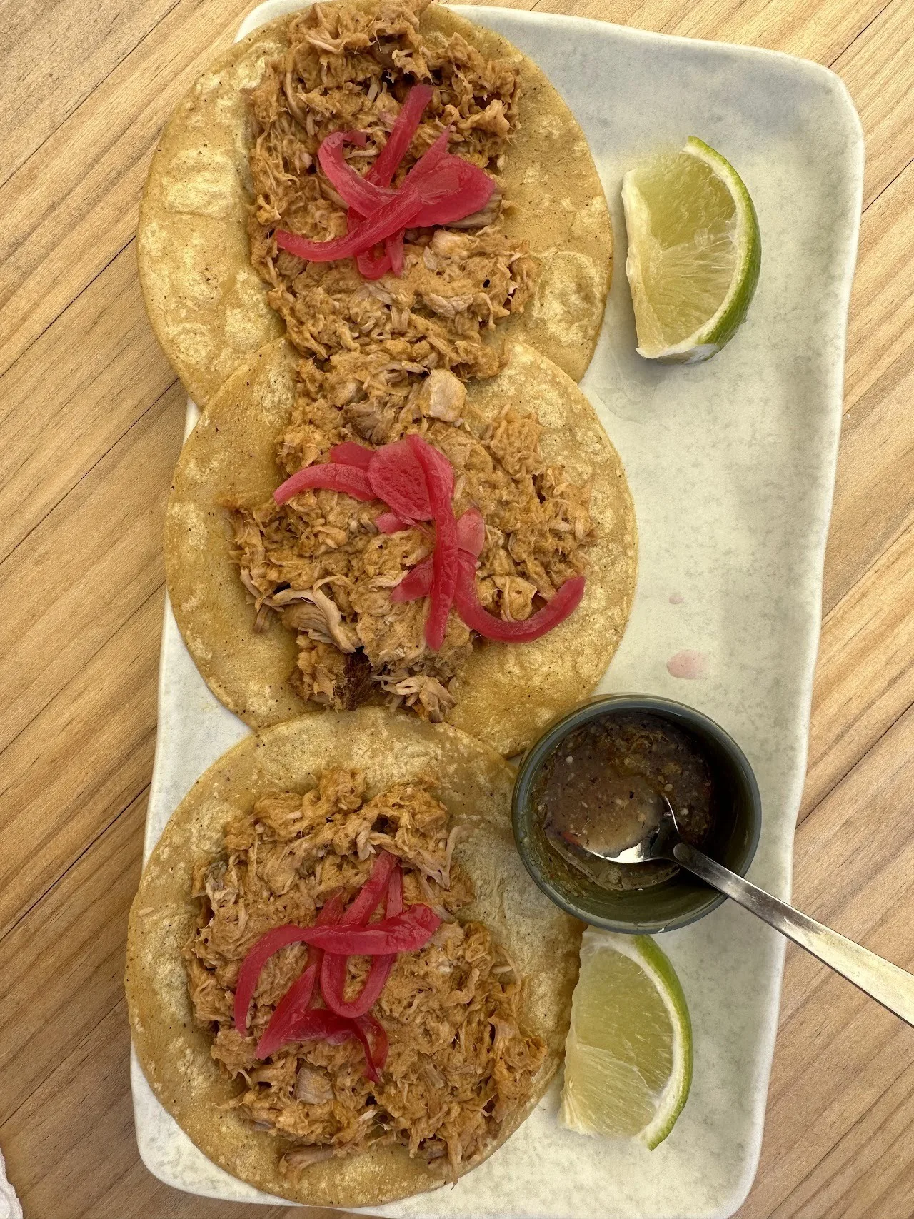 Three tostadas topped with shredded meat and pink pickled onion on a long plate with lime and a small dish of sauce
