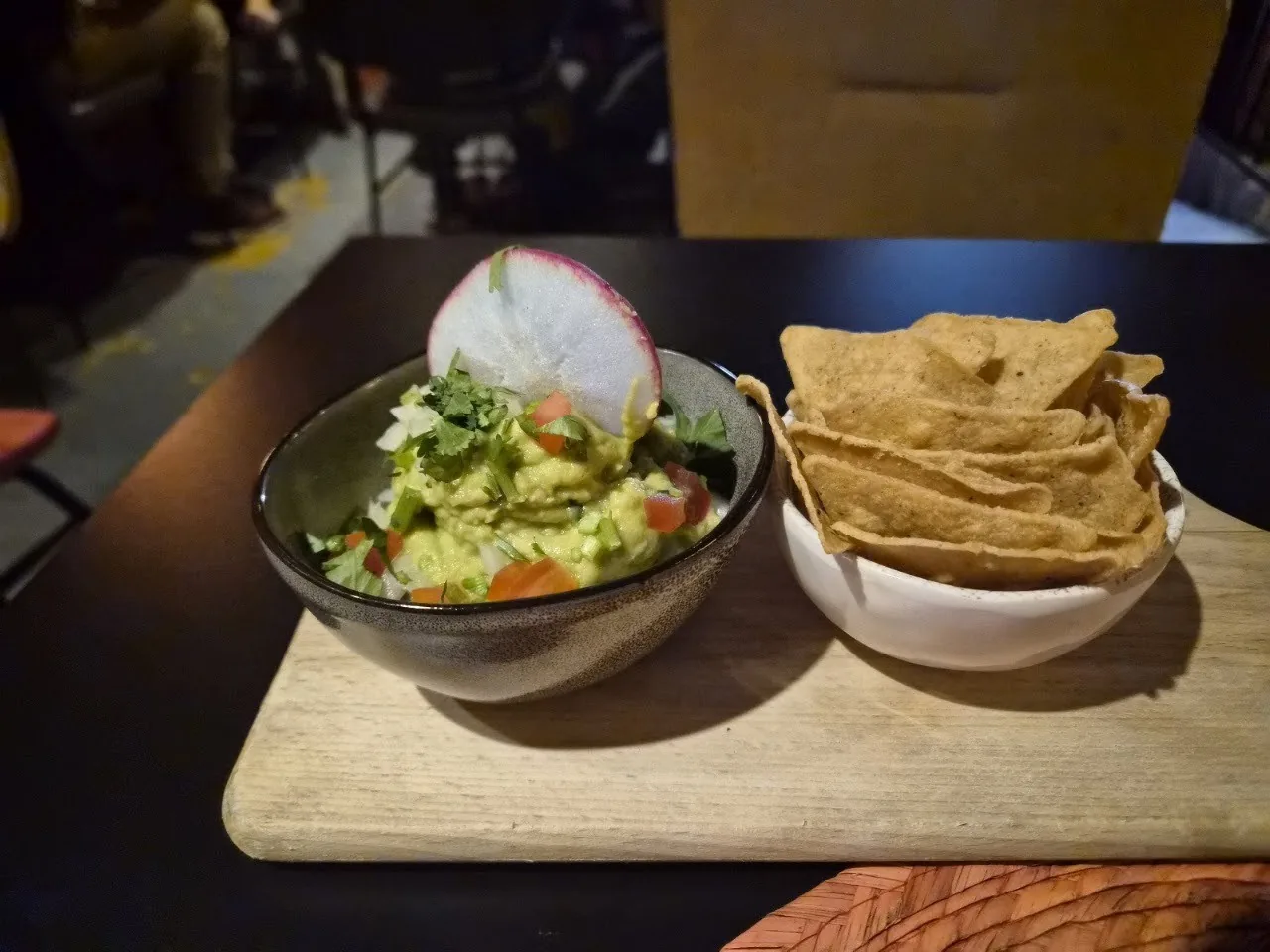 A bowl of guacamole with a radish slice, next to a bowl of tortilla chips on a wooden board
