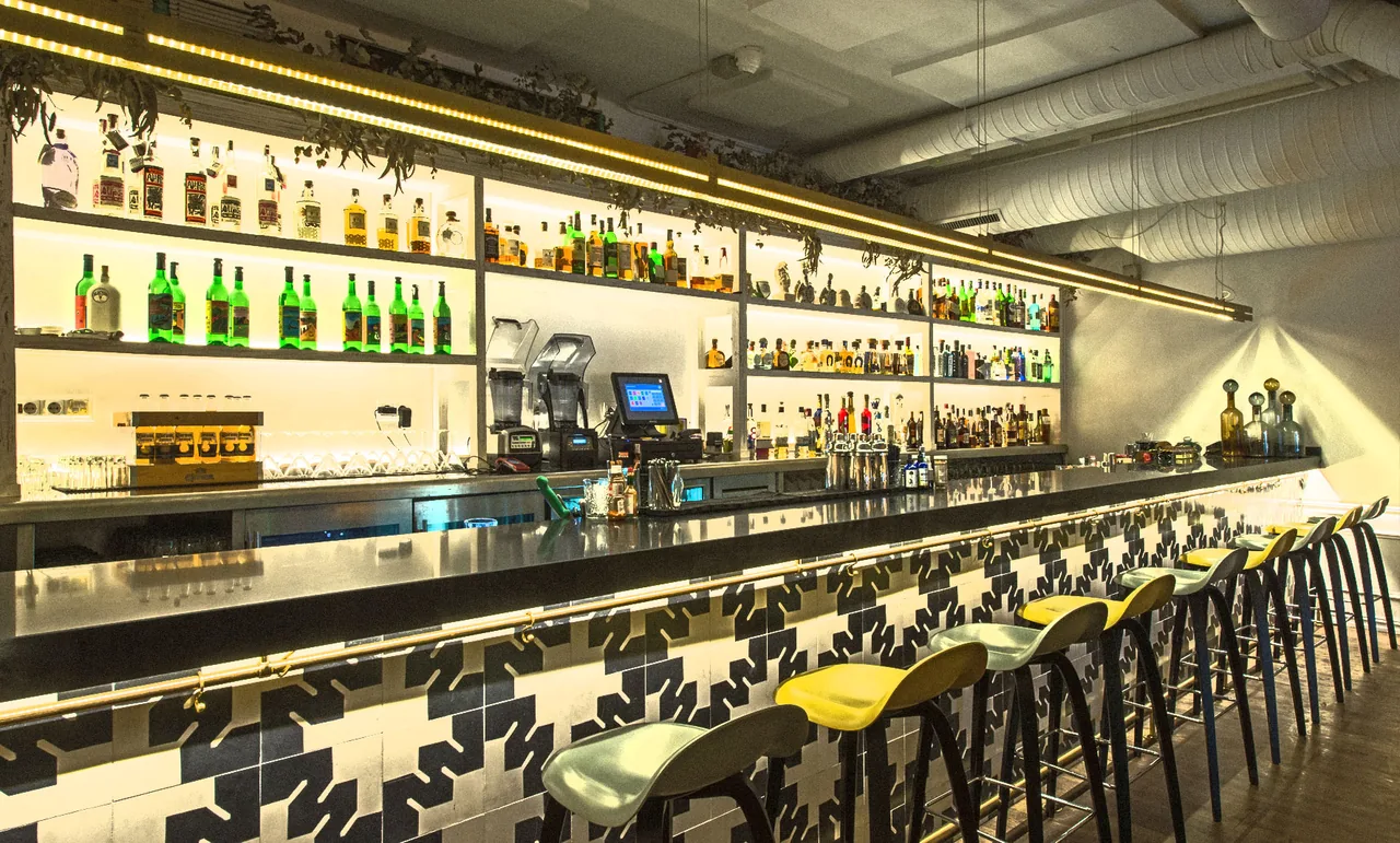 Long bar counter with a black and white patterned tile front and a row of yellow and pale green stools, backed by backlit shelves of agave spirits and mixers under strip lighting