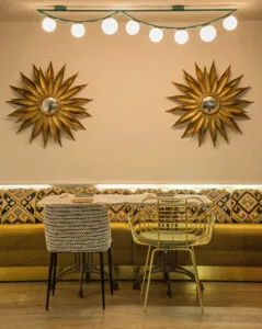Dining nook with two gold sunburst mirrors on a pink wall above a patterned banquette, small round tables with woven and gold chairs, under a string of globe lights