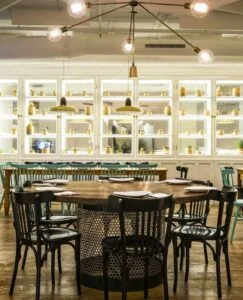 Dining room of round wooden tables and dark bentwood chairs in front of tall backlit white display shelving, lit by a branching pendant fixture of bare bulbs