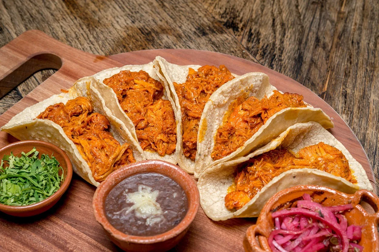 A wooden board of folded tacos filled with orange shredded meat beside bowls of beans and pickled onion