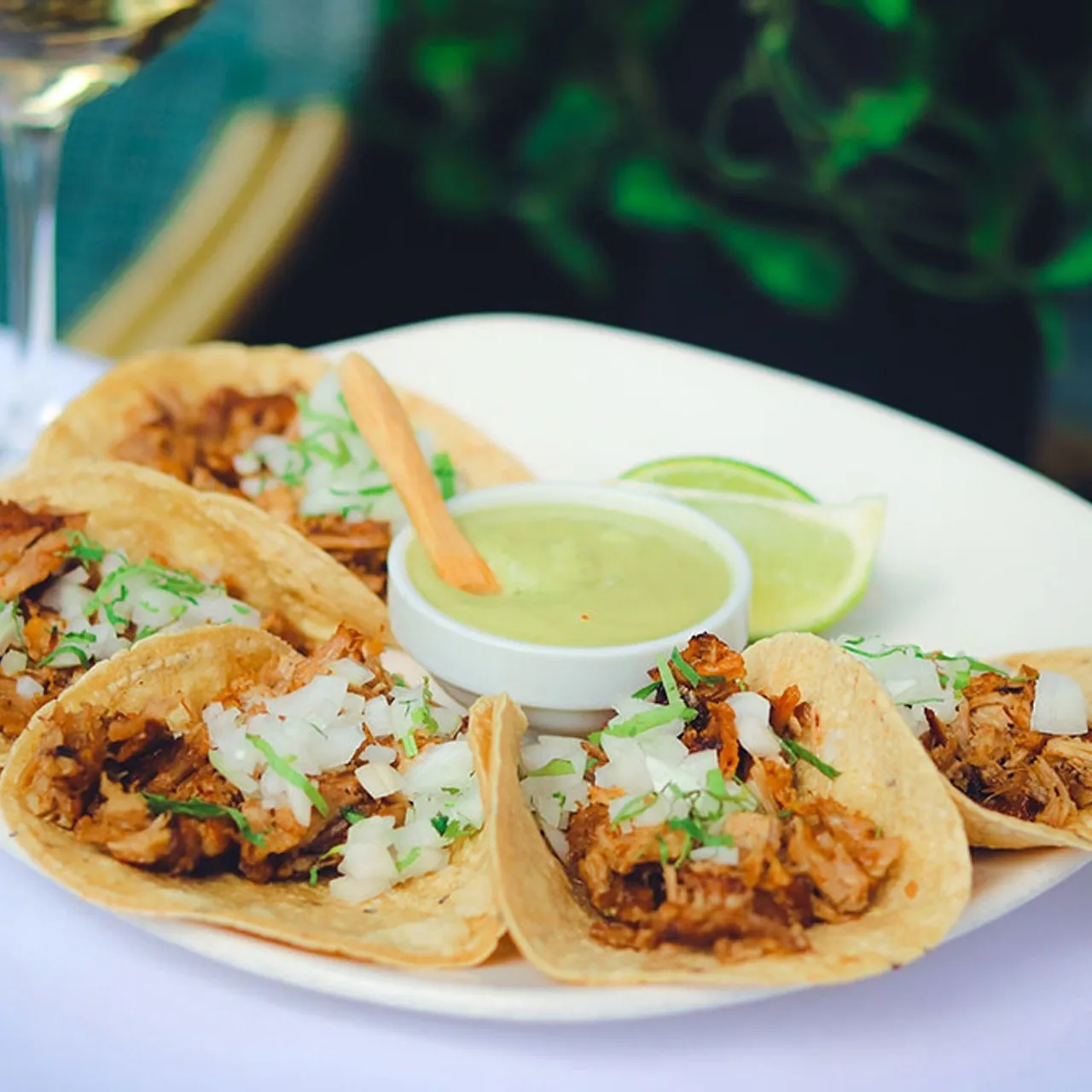 A plate of small tacos with chopped onion and herbs, a lime wedge and a bowl of green sauce