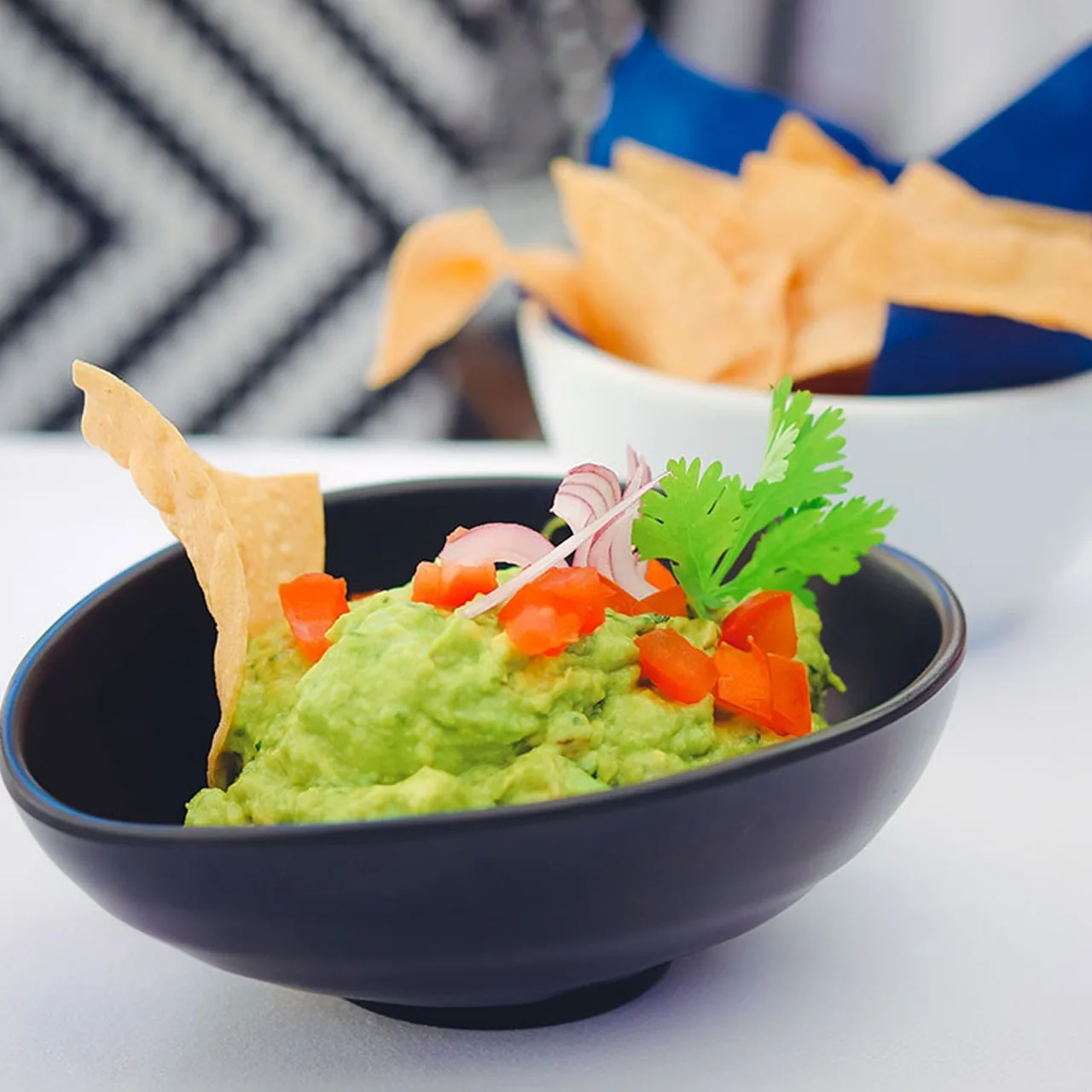 A dark bowl of guacamole with diced tomato and herbs, with tortilla chips behind