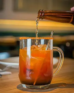 A drink being poured from a bottle into a glass mug filled with ice and orange liquid.