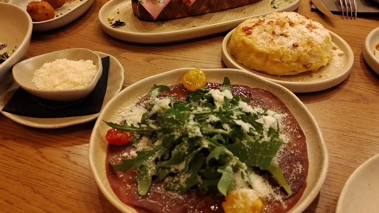 Beef carpaccio topped with arugula and shaved cheese, plated alongside other small dishes.
