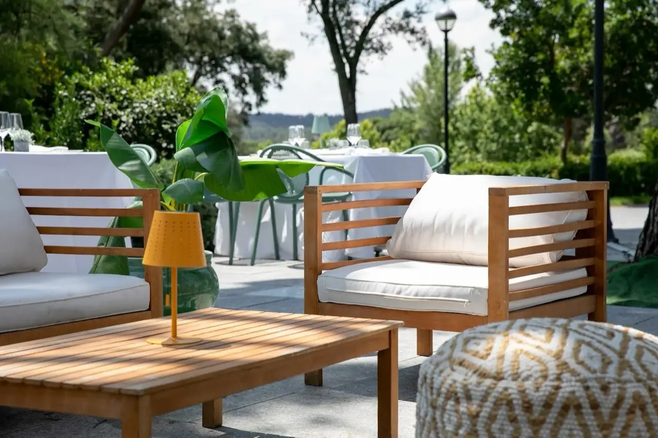 Outdoor restaurant terrace lounge with wooden armchairs and cushions, a slatted coffee table with a yellow lamp, and white-clothed dining tables amid greenery