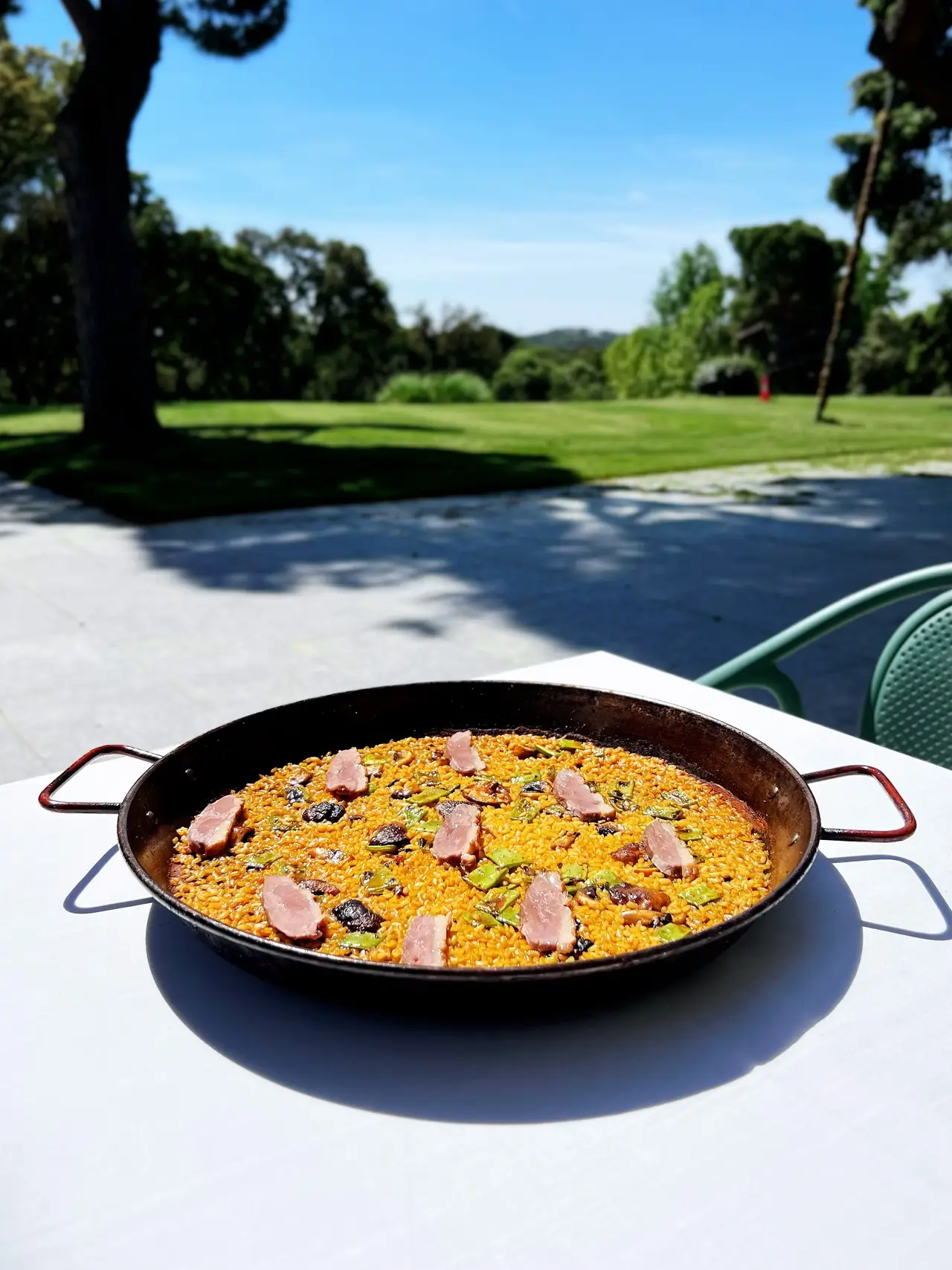 A paella pan of golden rice topped with slices of pink meat, on a white table on the terrace with the golf course behind