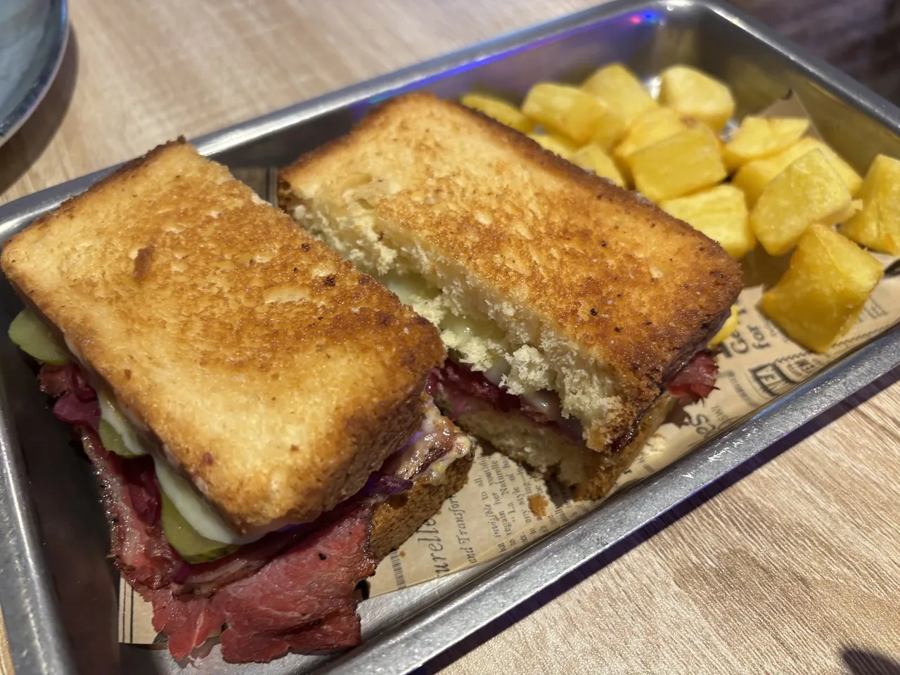 A toasted sandwich with sliced cured meat cut in half, served in a metal tray with fried potato cubes