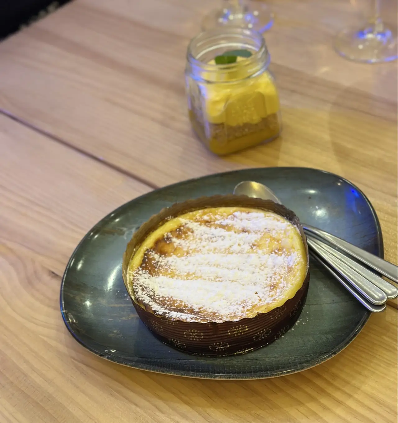 Round baked tart dusted with powdered sugar on a plate, with a jarred dessert in the background