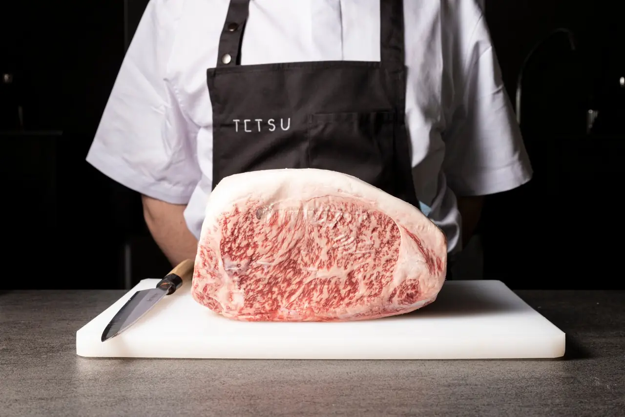 A heavily marbled whole beef striploin with a thick fat cap resting on a white cutting board beside a Japanese knife, held by a cook in a TETSU apron