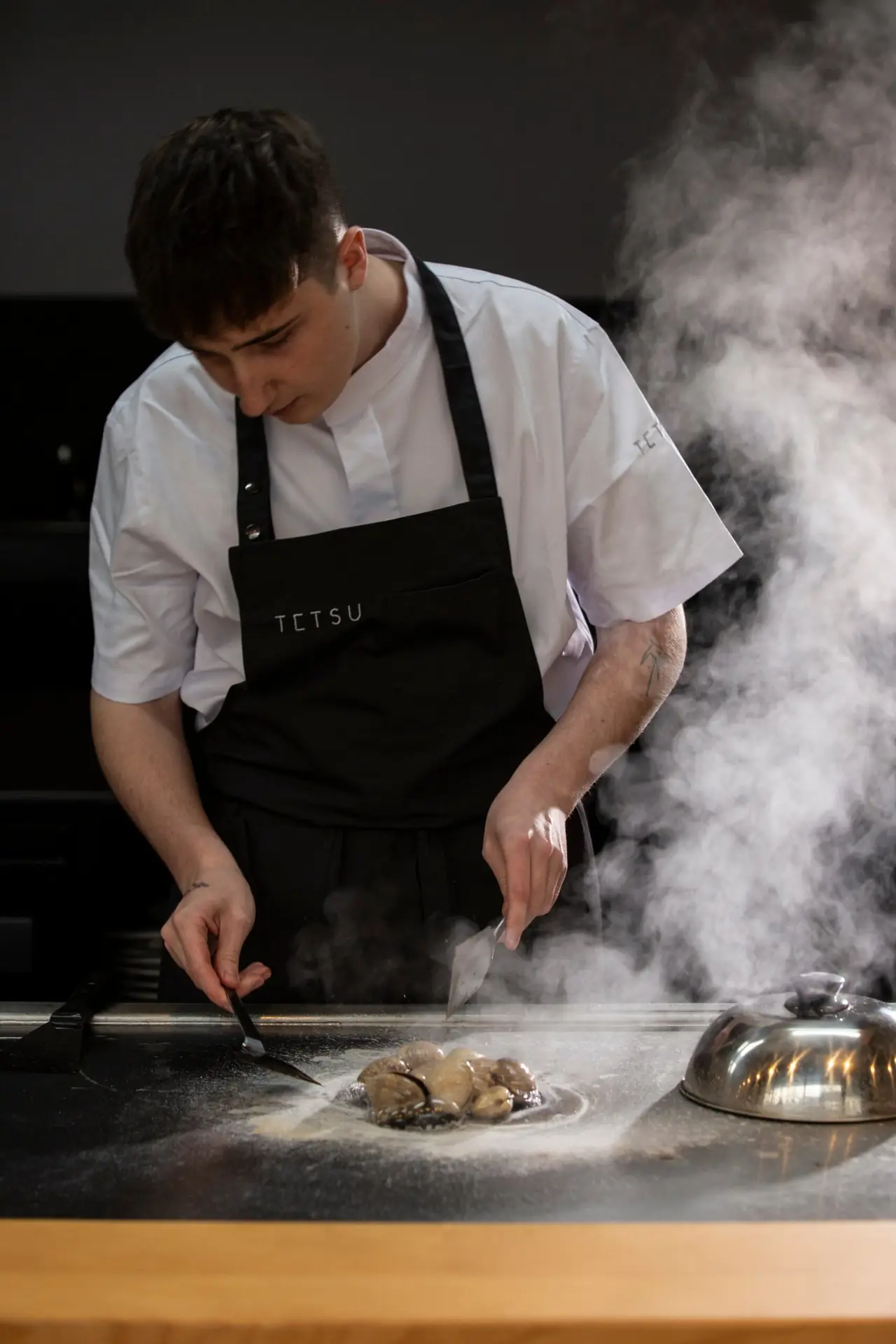 Chef working shellfish on a smoking teppan grill with two spatulas