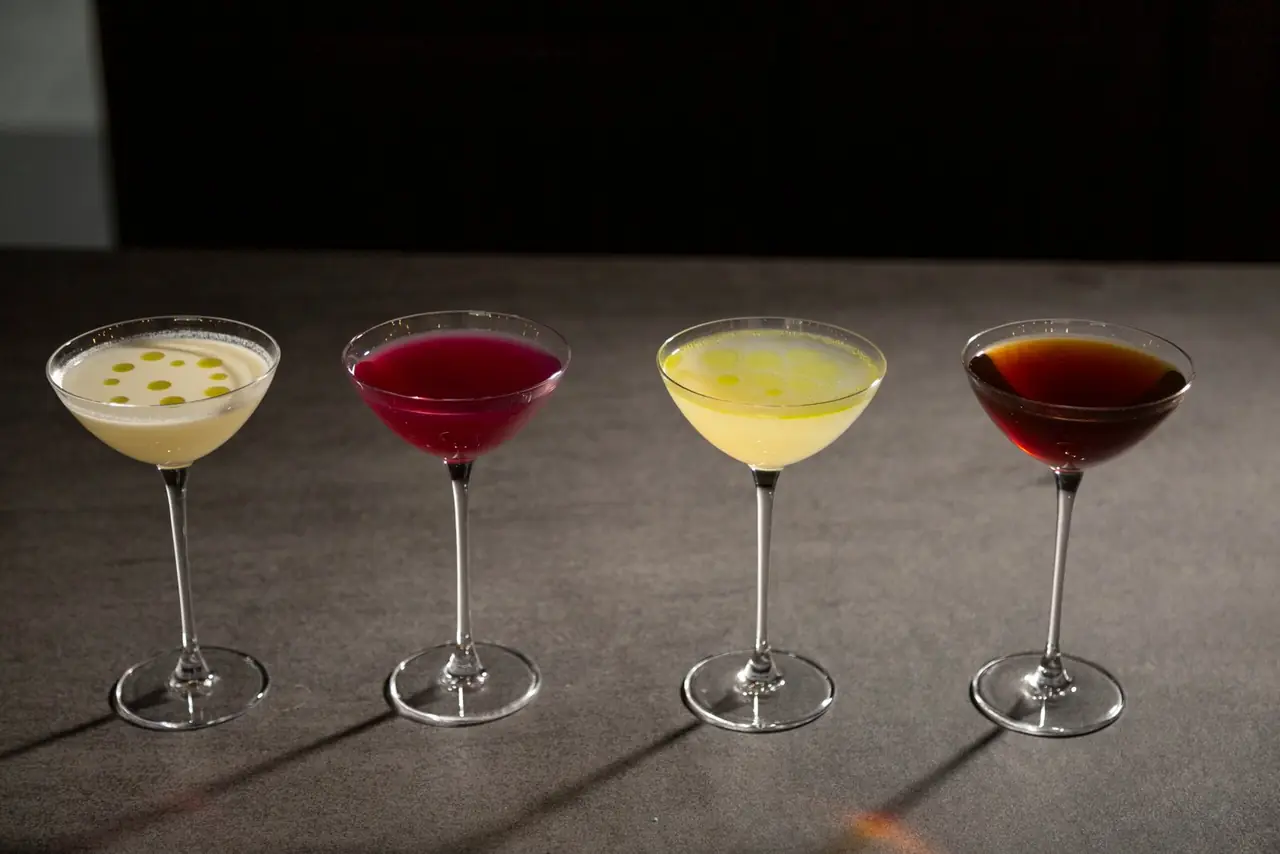 Four cocktails in coupe glasses in a row, cream, magenta, yellow and dark amber