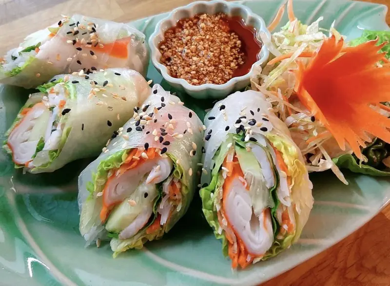 Fresh rice paper rolls filled with vegetables and shrimp, garnished with sesame seeds, next to a bowl of dipping sauce, Thai Zaap