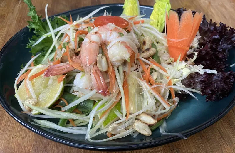 Green papaya salad with shrimp, peanuts, lime wedges, and mixed lettuce on a dark plate, Thai Zaap
