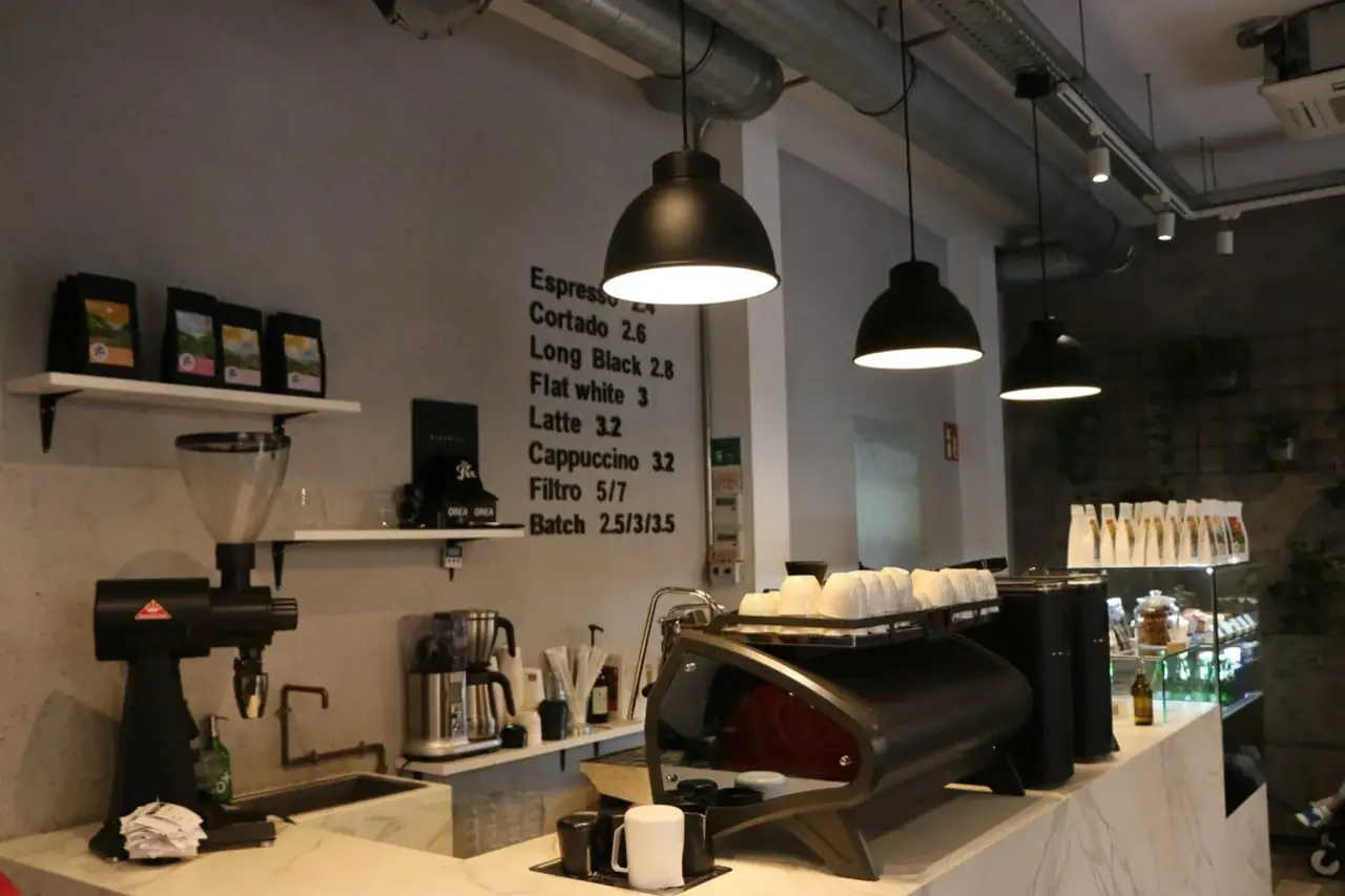 Cafe counter interior with an espresso machine, grinder, coffee bags on a shelf, hanging black pendant lamps and a handwritten price menu on the concrete wall