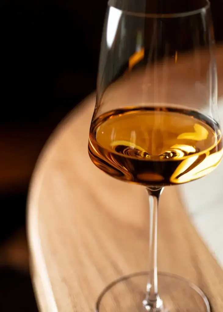 Stemmed glass of amber sherry or wine on a curved wooden table lit against a dark background