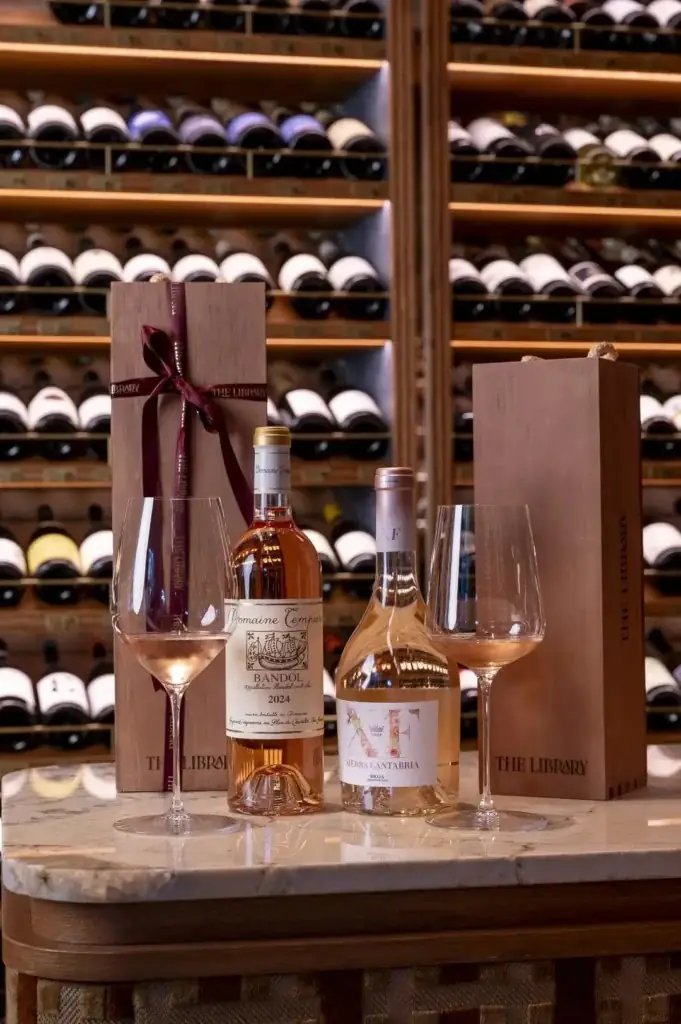 Two rose wine bottles with poured glasses and The Library branded wooden gift boxes on a marble bar in front of a wine cellar