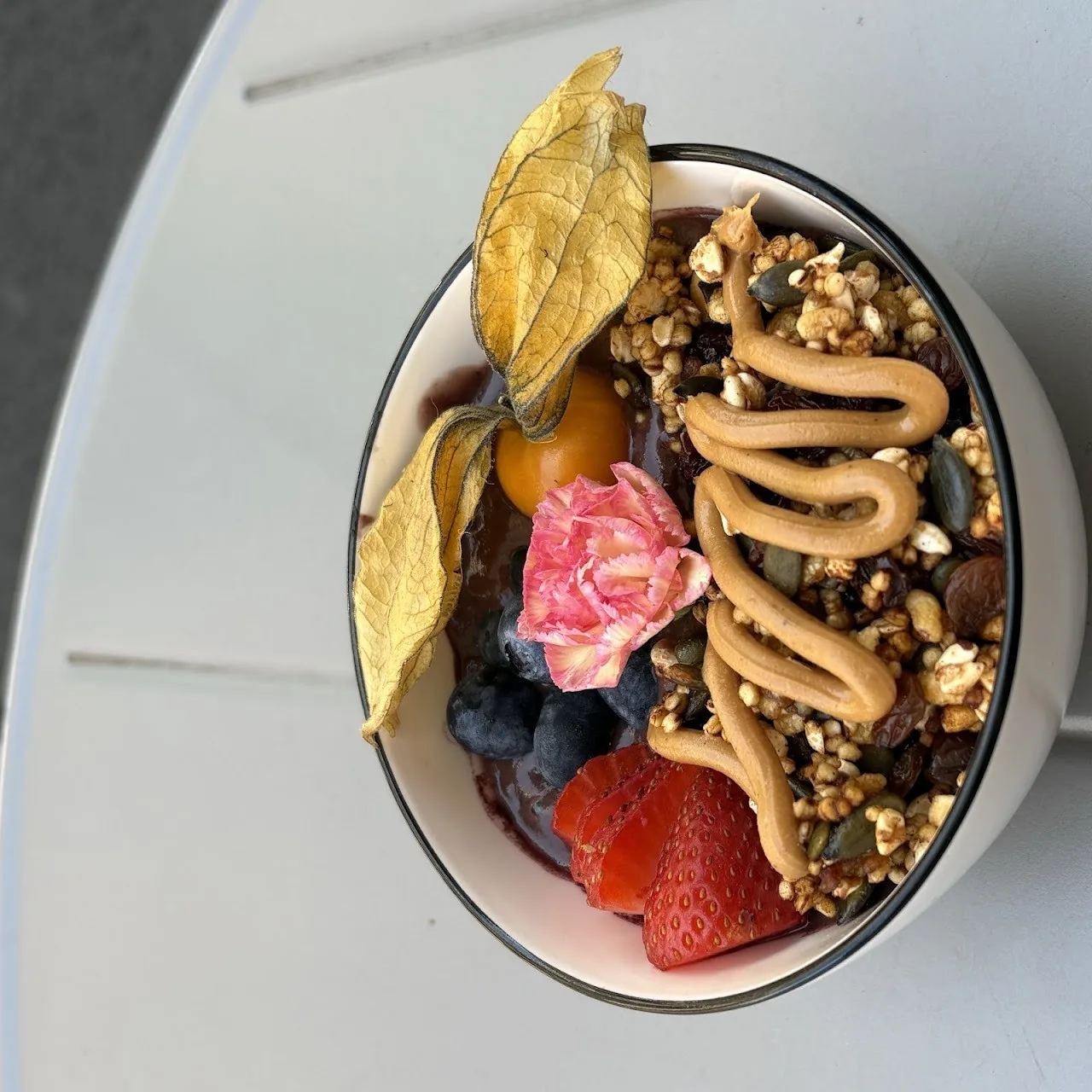 A bowl of granola with peanut butter, strawberries, physalis and an edible flower