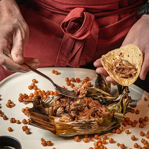 Hands lifting shredded braised meat with a fork from a charred banana-leaf parcel on a white plate and loading it onto a folded tortilla, crisp golden pearls scattered around