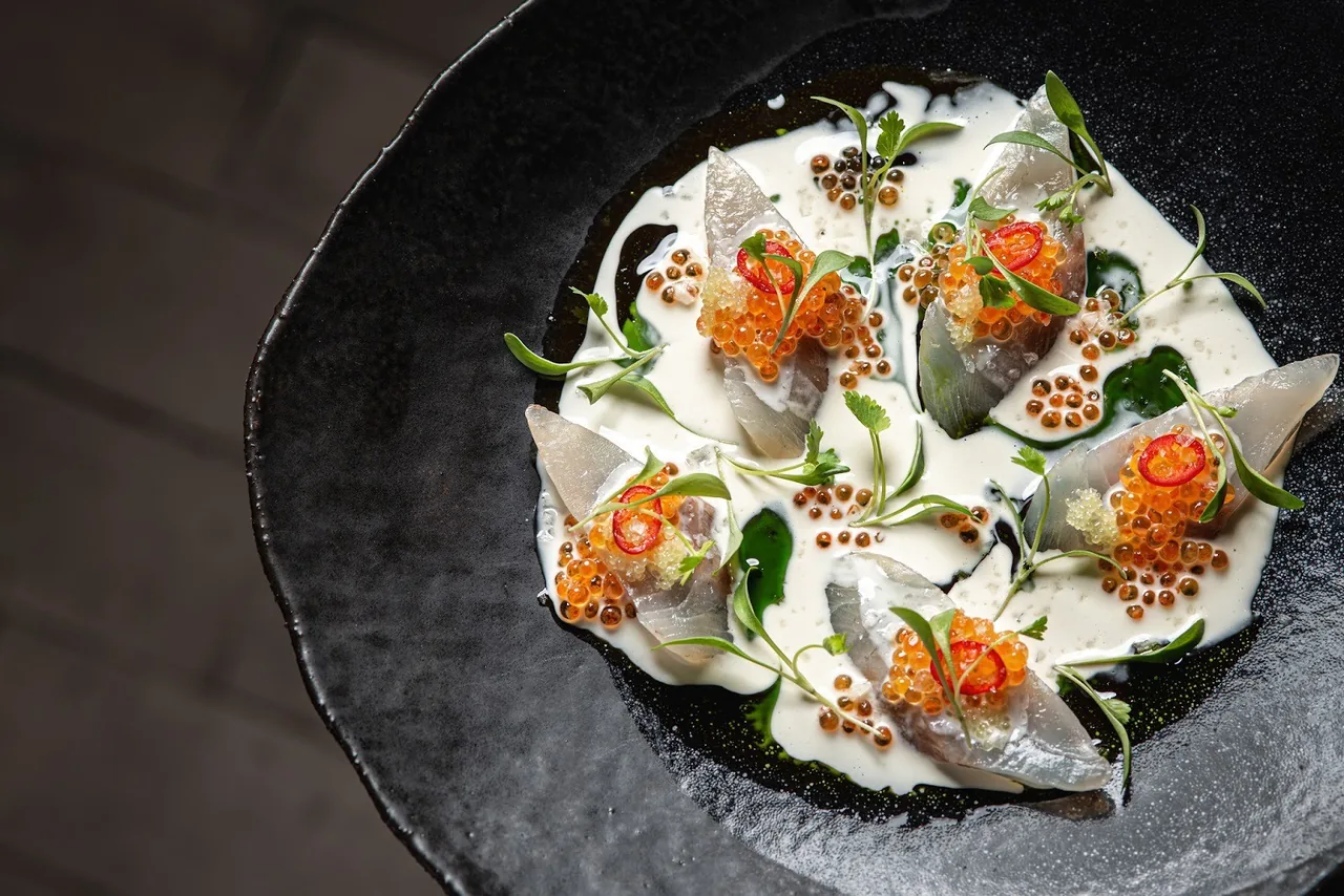 A dark plate of white cream topped with orange roe, herbs and small dabs of sauce