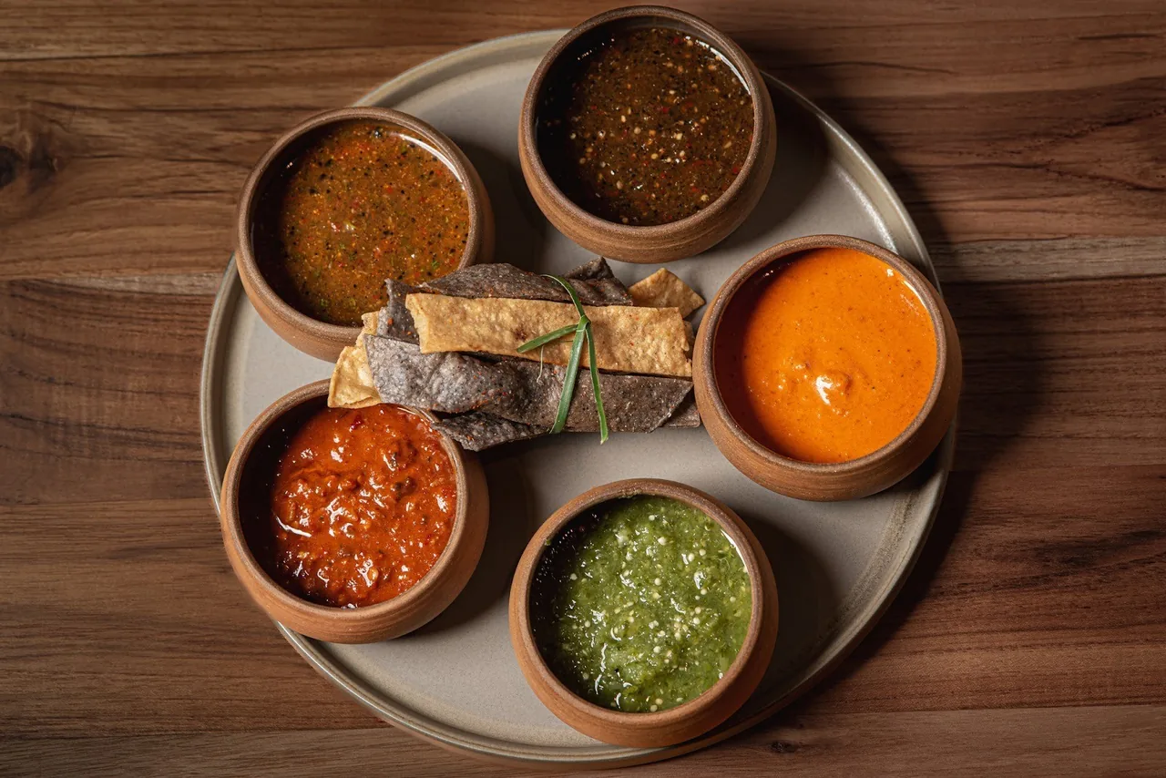 Five small clay bowls of different coloured salsas on a round plate with tortilla chips