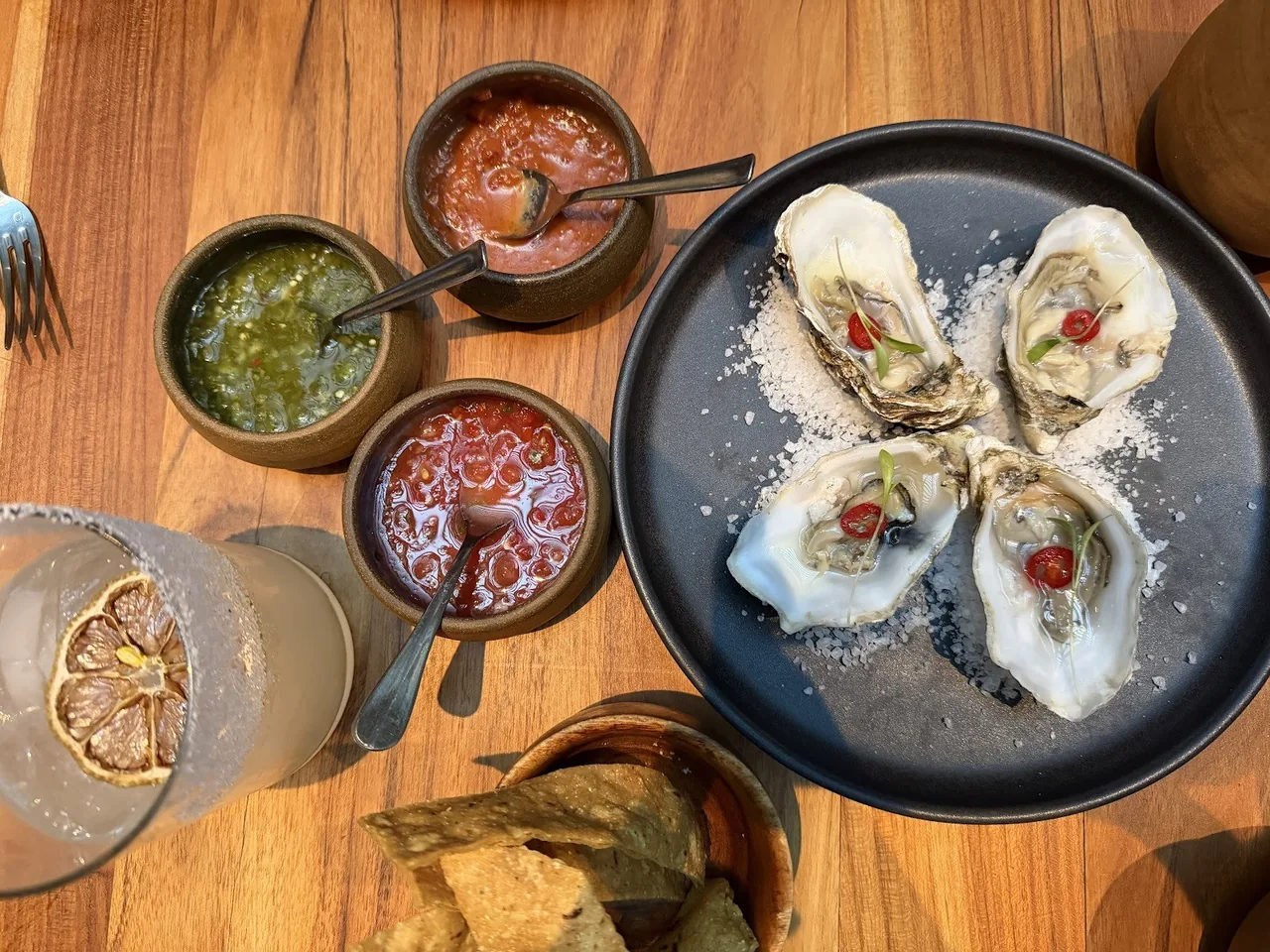 Four oysters on salt beside three bowls of salsa and a bowl of tortilla chips on a wooden table