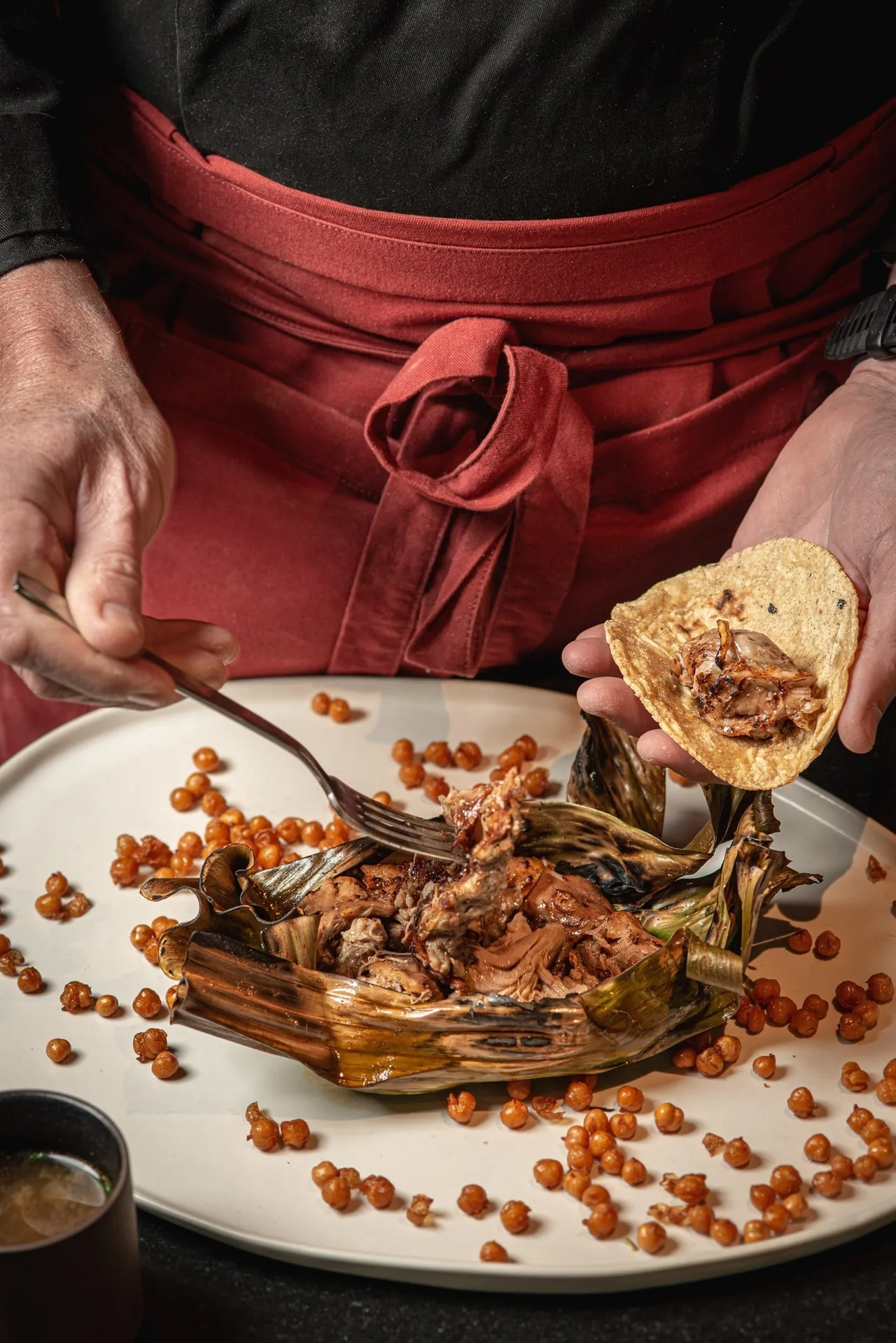 Shredded meat on a plate with scattered chickpeas while a hand fills a small tortilla