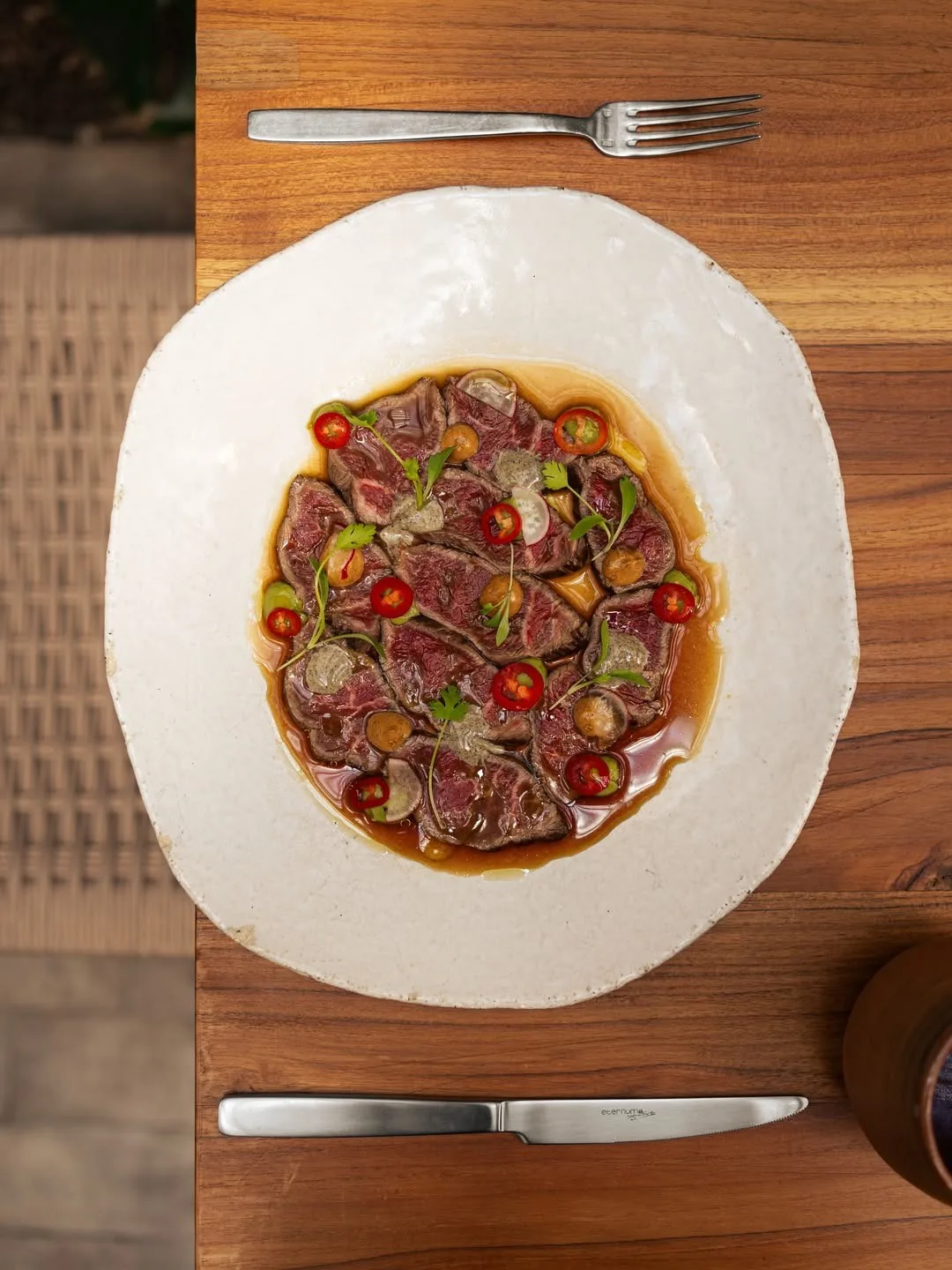An overhead view of a round white plate of sliced raw meat with red peppers and herbs on a wooden table