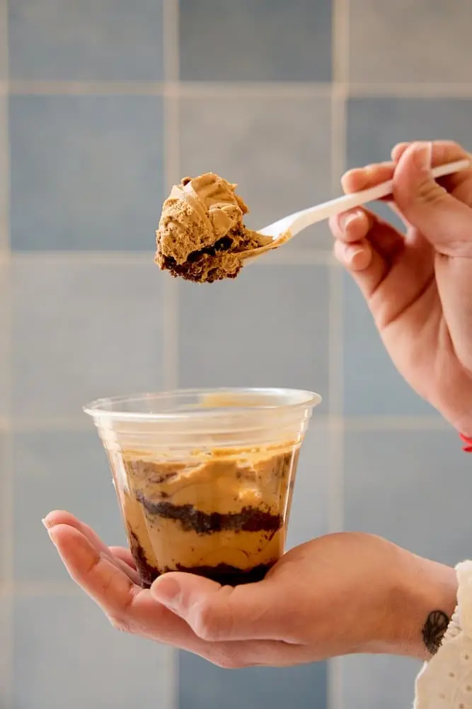 Hands holding a clear cup of layered chocolate and dulce de leche dessert with a spoonful lifted above