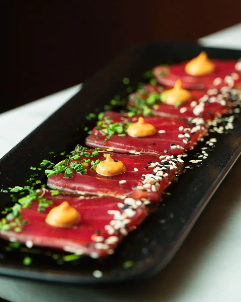 Row of tuna sashimi slices topped with dots of spicy orange mayo, chopped chives and black-and-white sesame seeds on a long black plate