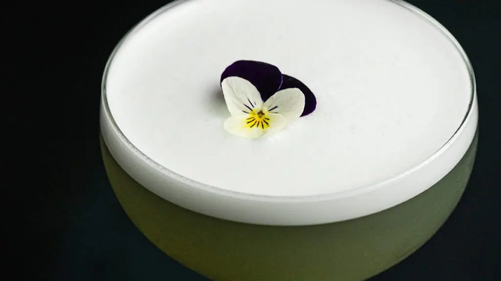 Frothy white-topped sour cocktail garnished with an edible white-and-purple pansy flower in a coupe on a dark surface