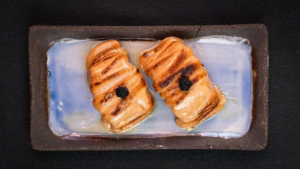 Two torched, seared nigiri-style pieces glazed in sauce with a black garnish, plated on a blue-glazed ceramic dish