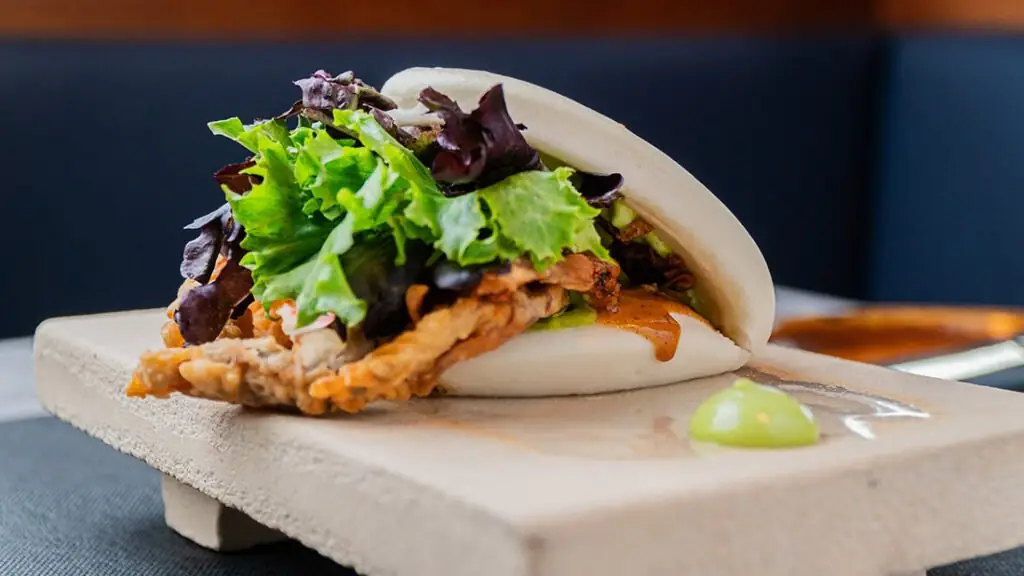 Bao bun filled with fried protein and mixed salad greens, served on a stone board with a dot of wasabi