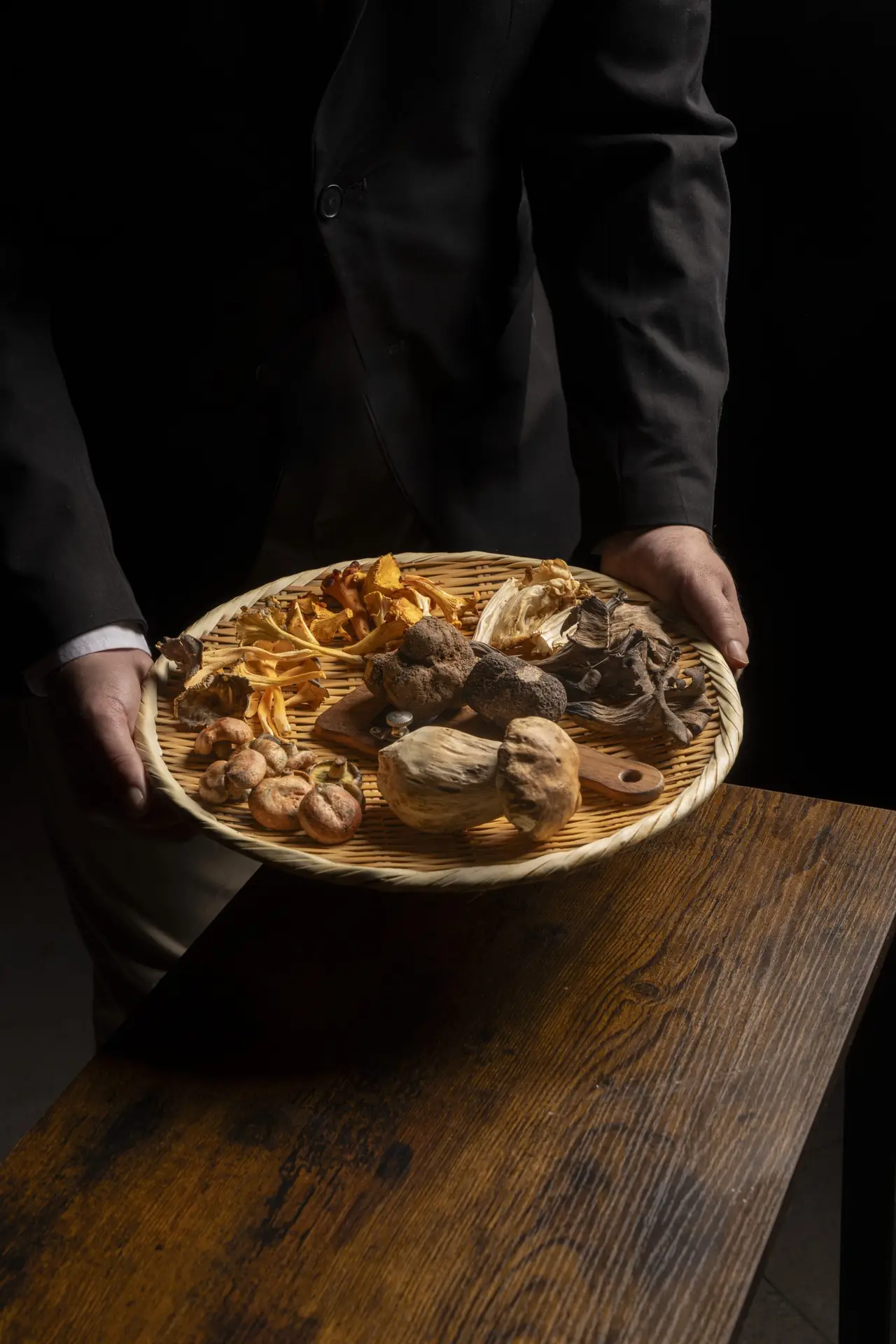 Server in black holding a woven basket of wild mushrooms and truffles above a wooden counter.