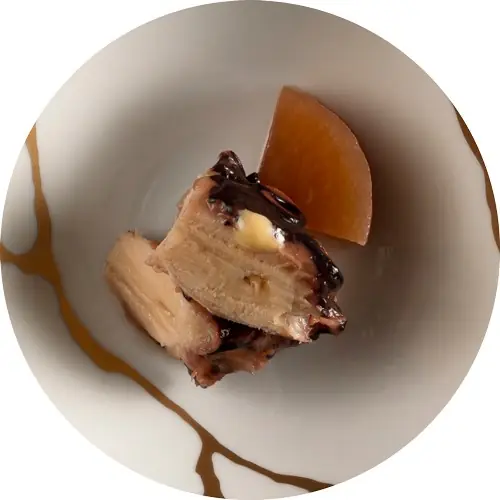 Circular crop of a pale braised dish with a wedge of simmered daikon and a dab of mustard on a marbled plate.