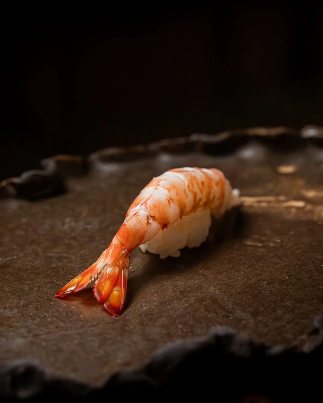 A single prawn nigiri on a dark rustic plate