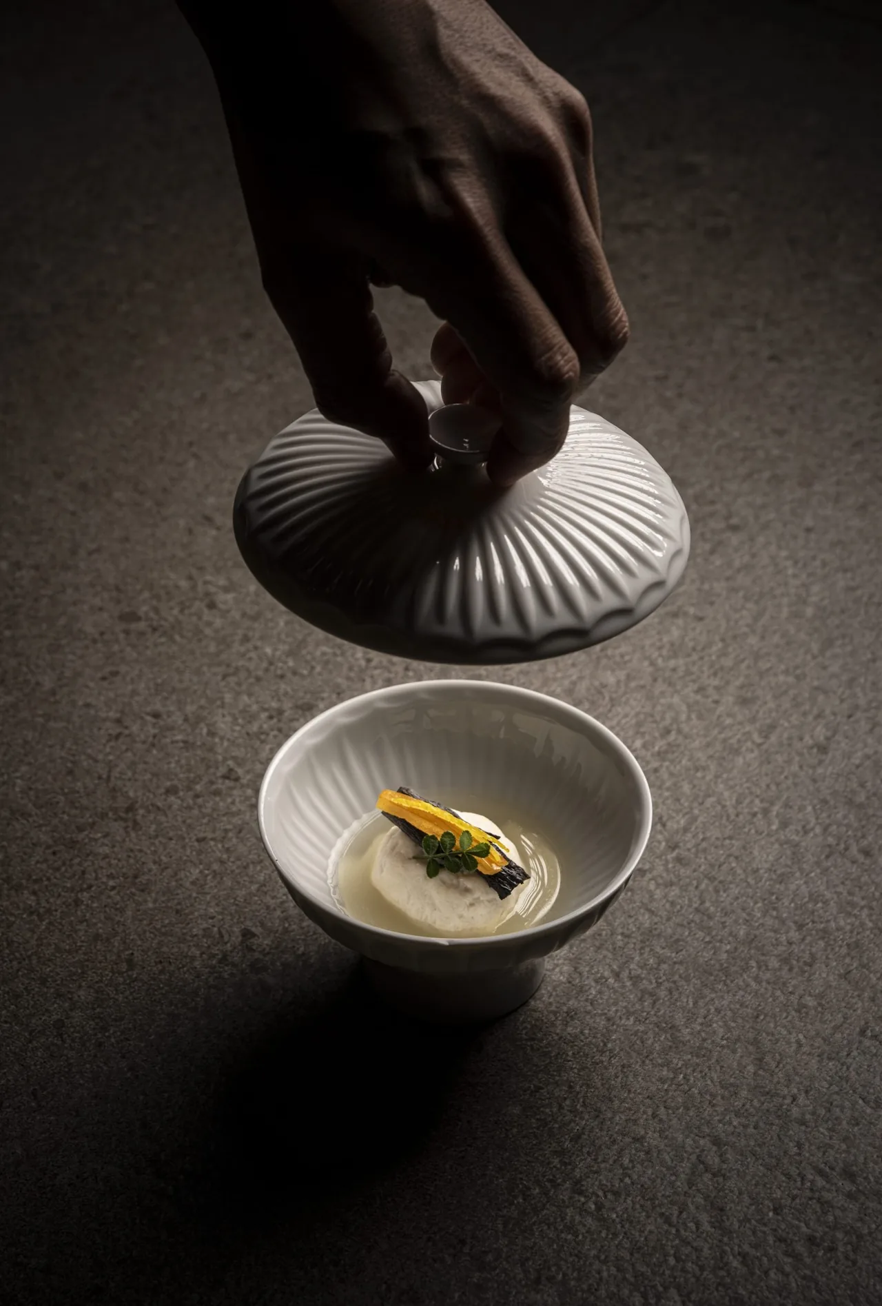 A hand lifting a ridged porcelain lid from a small bowl of pale custard in clear broth
