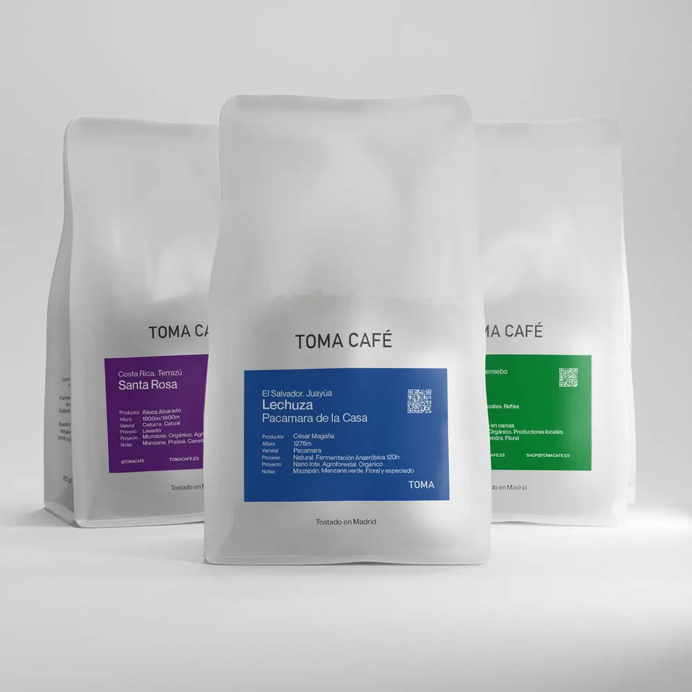 Three Toma Café coffee bags (Santa Rosa, Lechuza, and a green-labelled roast) standing on a plain studio background, retail product shot