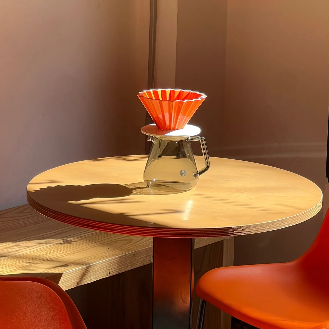 An orange pour-over dripper on a glass server sitting on a round wooden table in sunlight