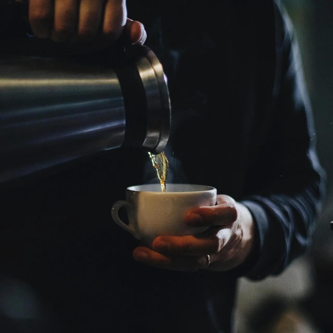 A hand holding a white cup while a stream of coffee is poured into it from a kettle